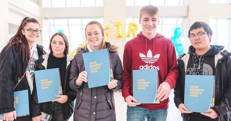 A group of five students holding light blue folders that say,