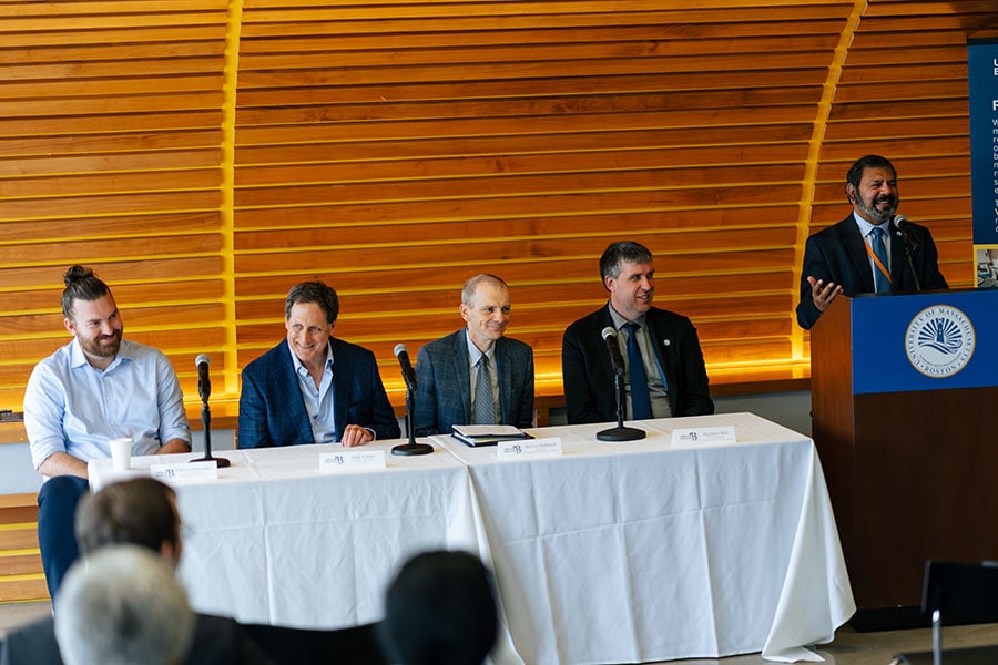 Bala Sundaram laughs at a podium, while the four panelists on the quantum roundtable sit at a table covered in a white tablecloth